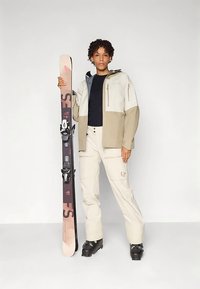 Άτομο με μπεζ και μαύρο εξοπλισμό σκι στέκεται κρατώντας ένα ροζ και μαύρο σκι όρθιο πίσω από έναν απλό φόντο.