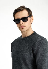 Gafas de sol negras con montura rectangular, llevadas por un modelo que viste un suéter gris texturizado con un diseño de canalé y cuello redondo.