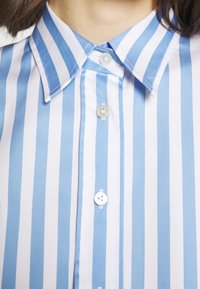 Gros plan d'une personne portant une chemise à rayures verticales bleues et blanches avec col, montrant trois boutons blancs boutonnés.