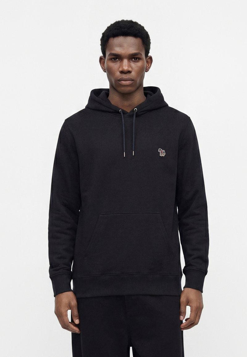 Jeune homme portant un sweat à capuche noir avec un petit logo animalier coloré sur la poitrine, debout devant un fond blanc uni.