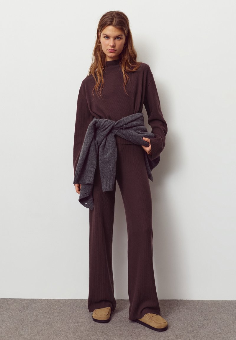 Bruine geribde trui en broek met wijde pijpen, gecombineerd met een grijze gebreide omslag om de taille en beige slippers, tegen een neutrale achtergrond.