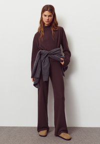 Pull marron côtelé et pantalon à jambes larges, associés à un cache-cœur en maille grise noué à la taille et des pantoufles marron clair, le tout sur un fond neutre.