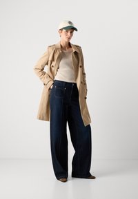 Trench beige sobre una blusa crema de canalé, jeans de pierna ancha azul oscuro y una gorra clara con acentos verdes. Zapatos marrones de punta.