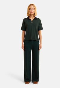 Dunkelgrünes Poloshirt mit schwarzem Kragen und halbem Reißverschluss, kombiniert mit dunkelgrünen weiten Hosen und braunen Plattformschuhen.