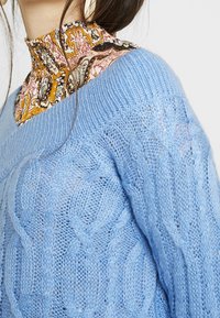 Suéter de punto trenzado azul claro con un escote amplio, superpuesto a una blusa de cuello alto con estampado que presenta colores cálidos y diseños intrincados.