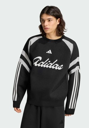 Junge Person mit schulterlangem dunklem Haar, die einen schwarzen Adidas-Pullover mit weißen Streifen und Logo trägt und vor einem hellen Hintergrund steht.