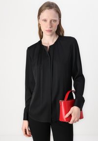 Svart langermet bluse med knappefeste og plisserte detaljer, kombinert med en liten rød lærveske med bærehåndtak.