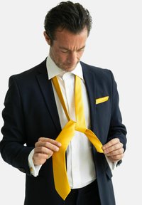 Gul silke slips lagt rundt halsen på en mand i en marineblå dragt, med en hvid skjorte og matchende lommetørklæde. Teksturfinish synlig.