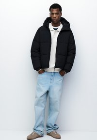 Sort pufferjakke med hætte, hvid skjorte med krave underneath, lyseblå løse jeans og beige sneakers. Enkel og funktionel design.