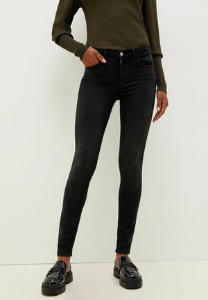 Vaqueros negros ajustados de mezclilla, con una cinturilla estándar, bolsillos frontales y una textura suave, acompañados de zapatos negros brillantes.