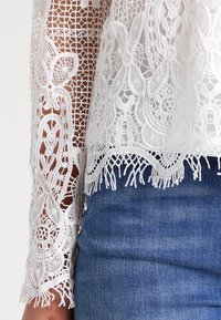 Top bianco in pizzo con motivi floreali intricati e orlo frangiato, dotato di maniche lunghe semitrasparenti, indossato sopra un denim blu.