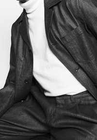 Chaqueta negra texturizada sobre un suéter de cuello alto blanco, con un bolsillo en el pecho y cierre de botones. Los pantalones de traje combinan con la tela de la chaqueta.