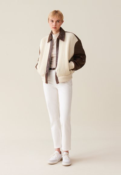 Oversized bomber bunda v krémovo-hnedej koži, s rebrovou textúrou, širokým golierom a rebrovanými manžetami, štýlovaná s bielymi nohavicami a teniskami.