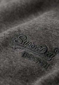 Tela gris con textura suave que presenta un logo "SuperDry" en relieve en un tono de gris más oscuro, mostrando un diseño simple y minimalista.