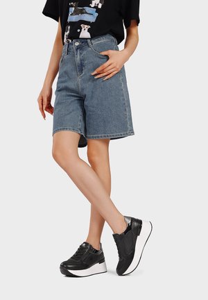 Persona che indossa pantaloncini di jeans a mezza lunghezza, una maglietta nera con grafiche di cani e sneakers nere con platform, in piedi con le gambe incrociate.