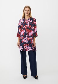 Blumige Tunika in Marineblau, Rot und Pink. Leichter Stoff, Knopfverschluss, Dreiviertelärmel, kombiniert mit weit geschnittenen, dunklen Denim-Hosen und schwarzen Schuhen.