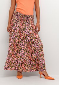 Jupe longue fleurie avec une taille smockée, ornée de fleurs roses et orange sur un fond vert. Associée à des escarpins à bride arrière orange.