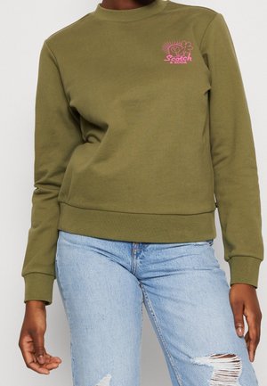 Persona con sudadera verde oliva con logo rosa en el pecho y vaqueros celestes rotos, de pie con las manos relajadas a los lados.