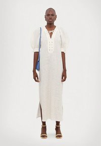 Moteris, vilkinti baltą midi suknelę su pūstomis rankovėmis, mažo gėlių rašto, su šoninėmis skeltukais, geltonais sandalais, auksinėmis auskarais-žiedais ir nešančia mėlyną rankinę ant peties.