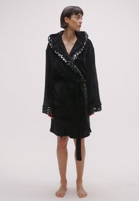 Albornoz de felpa negro con capucha adornada con lentejuelas, mangas largas, cintura ajustable con cinturón y bolsillos laterales. Textura suave y diseño hasta la rodilla.