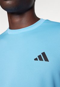 Svijetloplava atletska majica od teksturirane tkanine, s crnim adidas logotipom na lijevoj strani prsnog koša. Dizajn s okruglim izrezom.