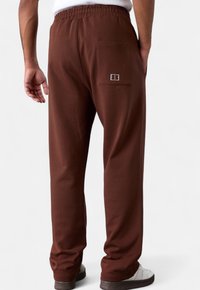 Braune Baumwollhosen mit einem elastischen Bund, ausgestattet mit einer Gesäßtasche und minimalen Nähdetails. Die abgebildeten Schuhe sind weiß mit braunen Sohlen.