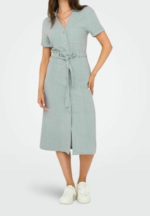 Žena ve světle modrých midi šatech s knoflíky na přední straně, krátkými rukávy, dvěma kapsami na prsou, zavazováním v pase a bílými teniskami.