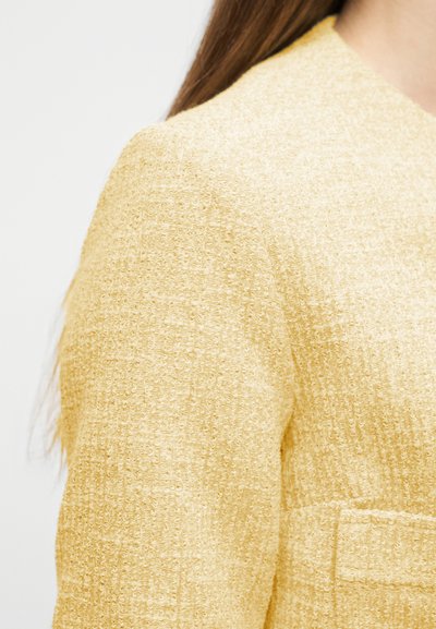 Gros plan sur l'épaule et le haut de la manche d'une veste en tweed jaune texturé portée par une personne aux longs cheveux bruns.