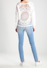 Top bianco a maniche lunghe con dettaglio in pizzo a uncinetto sulla schiena e motivi circolari, abbinato a jeans attillati azzurri e sneakers bianche.