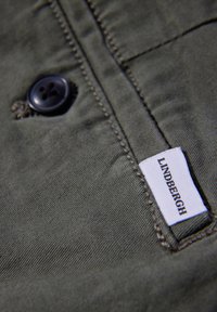 Tejido verde con una textura suave; presenta un botón oscuro, costuras visibles y una etiqueta blanca con el texto "LINDBERGH".