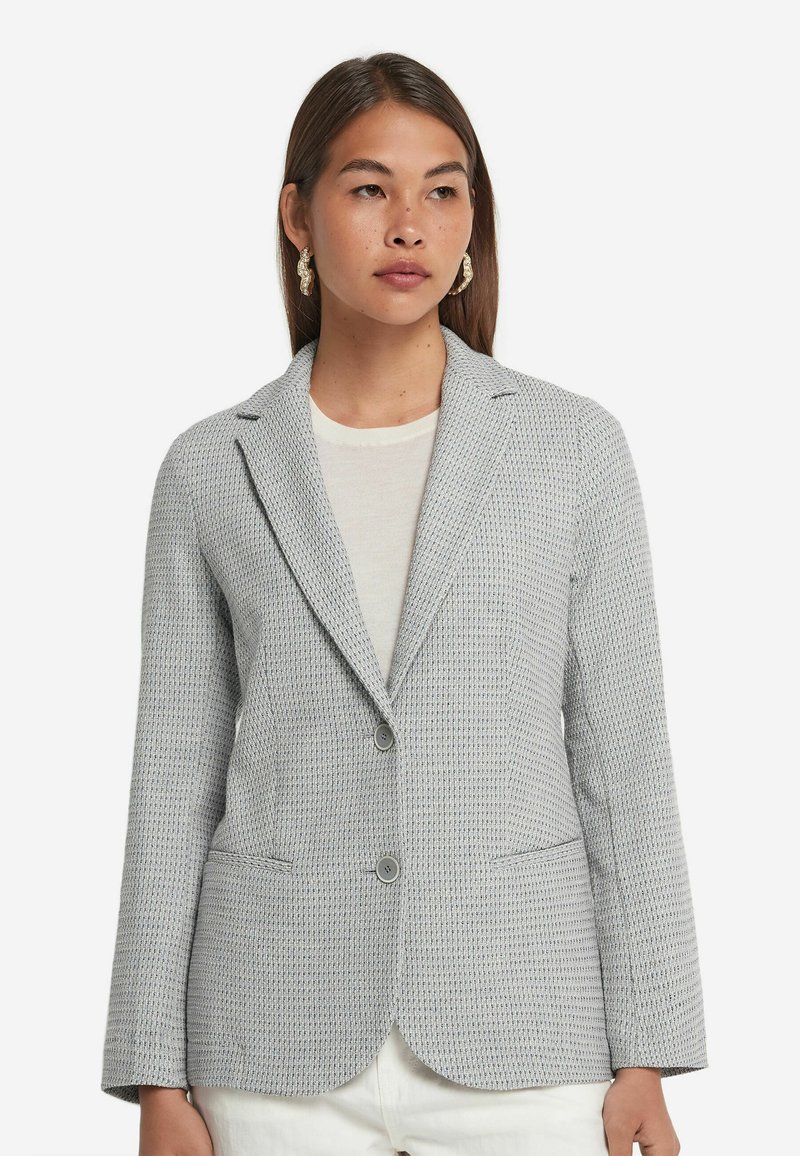 Grå texturerad kavaj med slag, två framfickor och två knappar. Slim fit-design, visad på en modell som bär en ljus topp.