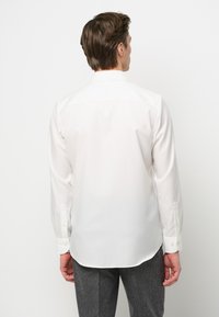 Camicia bianca a maniche lunghe con una texture liscia, polsini con bottoni e orlo curvo. Il retro presenta un colletto classico e dettagli minimi.