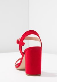 Red suede high-heeled sandal with a chunky heel, ankle strap, and open toe design. Smooth beige inner lining and adjustable buckle detail.