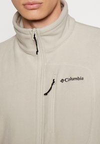 Ljusbeige fleecejacka med hög krage, dragkedja framtill, svarta detaljsömmar och en liten Columbia-logotyp på vänster bröst.