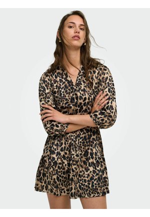 Femme aux longs cheveux bruns portant des boucles d’oreilles créoles en or et une robe à manches longues imprimée léopard, les bras croisés devant un fond clair.