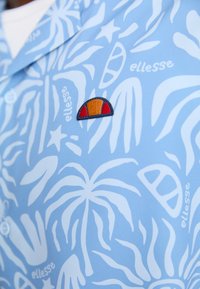 Lys blå skjorte med hvitt tropisk bladmønster, med et brodert oransje og rødt logo-patch på brystet. Knappelukking synlig.