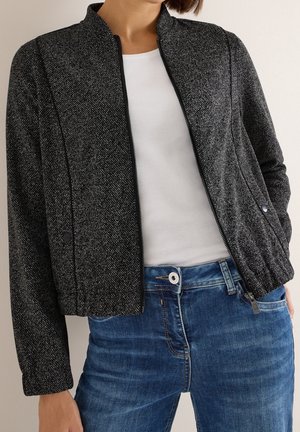 Femme portant une veste zippée noire texturée par-dessus une chemise blanche et un jean bleu, main dans la poche devant un fond uni.