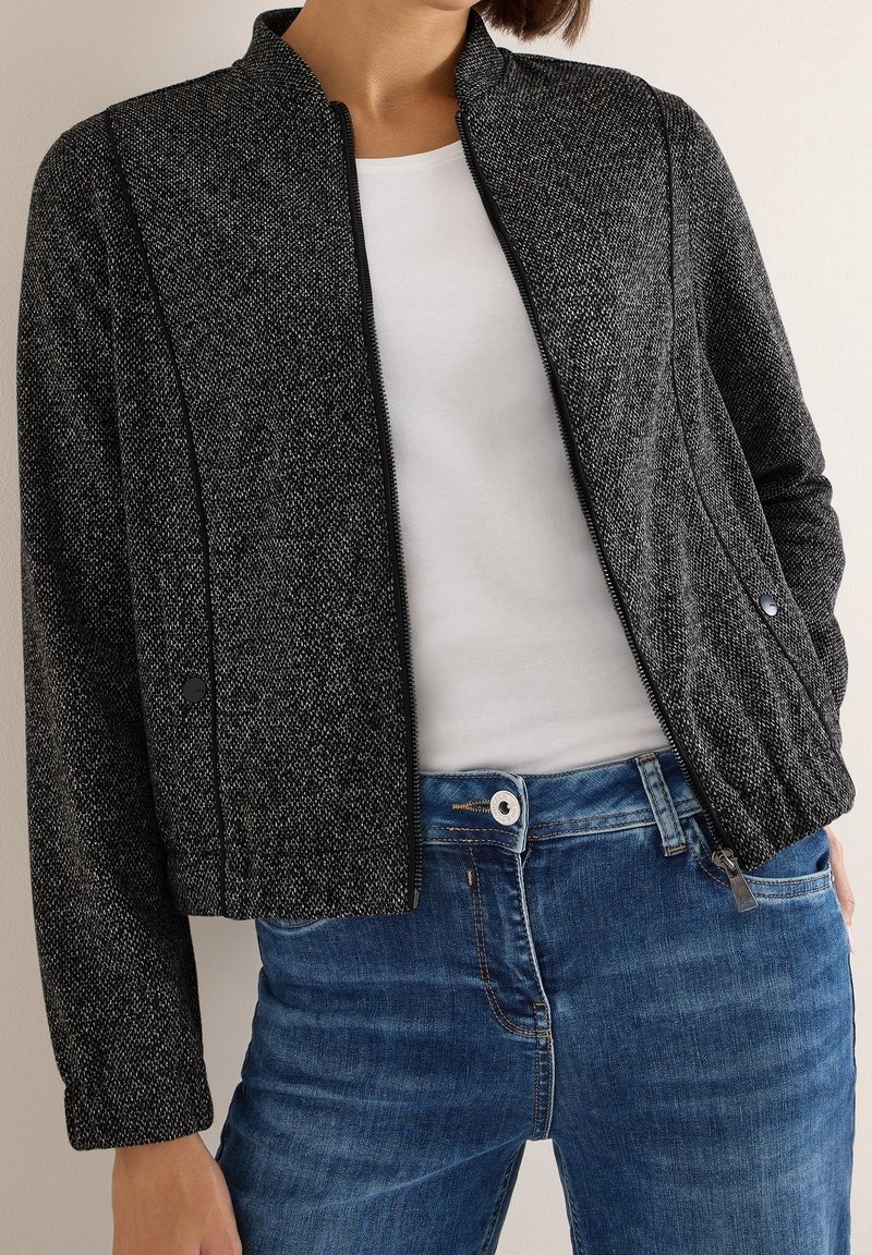 Femme portant une veste zippée noire texturée par-dessus une chemise blanche et un jean bleu, main dans la poche devant un fond uni.