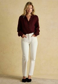 Burgundský kardigán s pleteným vzorom a tmavými gombíkmi, skombinovaný s bielymi džínsami s vysokým pásom a čiernymi chlpatými nazúvacími topánkami, na neutrálnom pozadí.