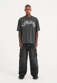 Camiseta gris de gran tamaño con "Ed Hardy" impreso en blanco. Combinada con pantalones cargo negros con múltiples bolsillos y detalles texturizados.
