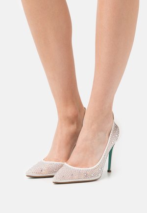 Pies de mujer luciendo tacones altos blancos y translúcidos con brillantes de strass y tacones de aguja verdes sobre un fondo blanco.