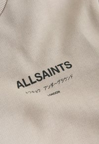 Gai beige audums ar tekstu "ALLSAINTS" treknā melnā fontā, zemākā līmenī papildus teksts mazākā fontā. Mīksta tekstūra, minimāls dizains.