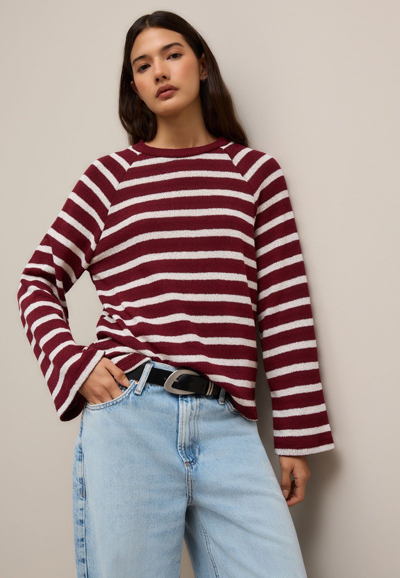 Maglione a maniche lunghe a righe bordeaux e bianche con collo rotondo e maniche a campana, abbinato a jeans azzurri a vita alta e cintura nera.