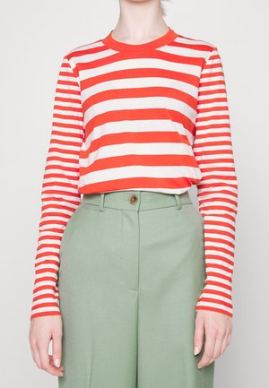 Personne portant une chemise à manches longues rayée horizontale rouge et blanche, rentrée dans un pantalon taille haute vert clair avec fermeture par bouton.