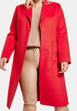 Vrouw draagt een felrode knielange jas over een beige gebreide trui en lichtbruine leren broek, staat met licht gespreide armen.