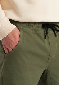 Olivgröna bomullsjoggers med elastisk midja, dragsko och sidofickor. Tyget verkar mjukt med en slät textur.