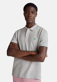 Šedé polo tričko s malým zeleným logem. Krátké rukávy a standardní límec. Model stojí s překříženýma rukama, má na sobě růžové šortky.