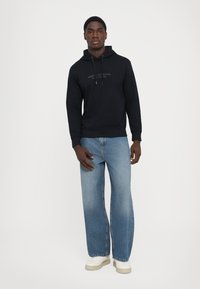 Sudadera con capucha de color azul marino con cordón, que presenta un logo en la parte frontal. Combinada con unos jeans de pierna ancha de color azul claro y zapatillas blancas. Textura suave.