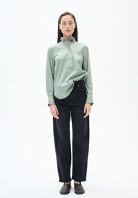 Hellgrüne Bluse mit Rüschensaum und halbem Knopfverschluss, kombiniert mit lockeren schwarzen Jeans und flachen dunkelbraunen Schuhen. Einfarbiger Hintergrund.