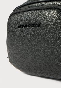 Čierna kožená taška s textúrou a zalisovaným logom "ARMANI EXCHANGE". Obsahuje zips na zatváranie a má hladký, odolný povrch.